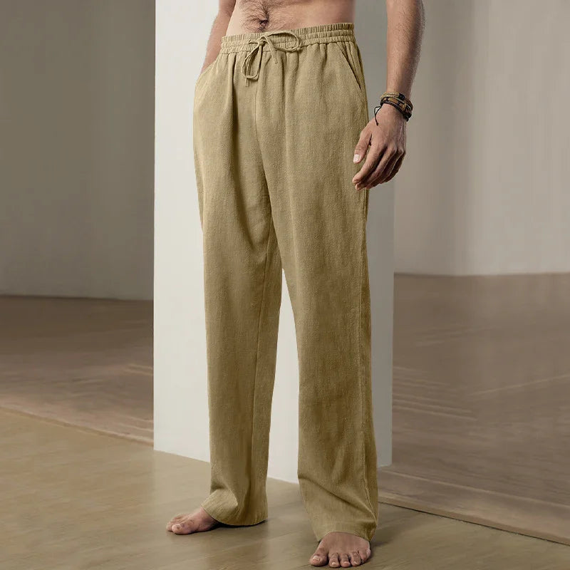 Man wearing beige linen drawstring pants standing barefoot indoors on wooden floor