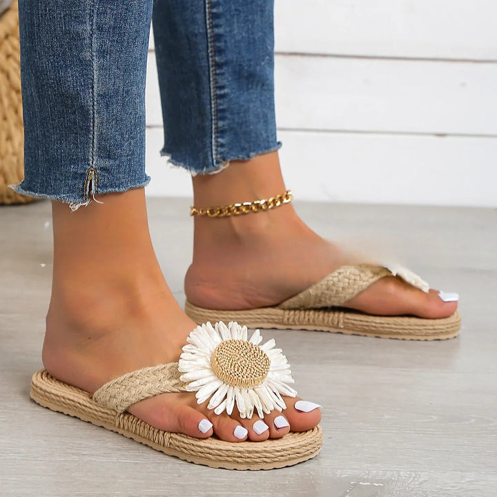 Feet wearing beige flip-flop sandals with large white daisy decoration and gold ankle chain, paired with frayed blue jeans