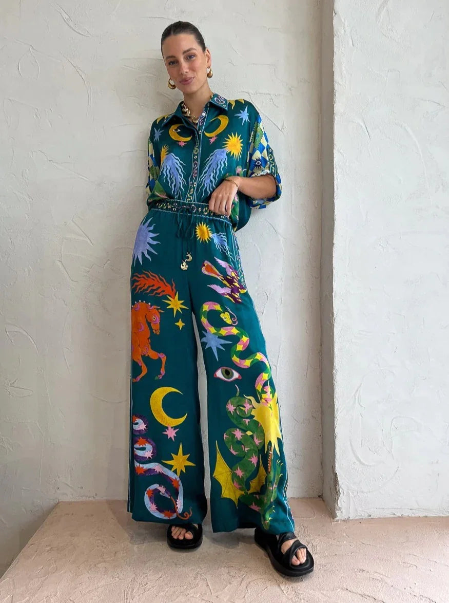 Woman in teal wide-leg pants and matching shirt with colorful celestial and animal prints standing against textured white wall