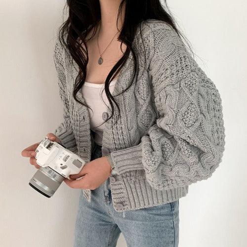 Woman wearing chunky gray cable knit cardigan and jeans holding white camera against white background