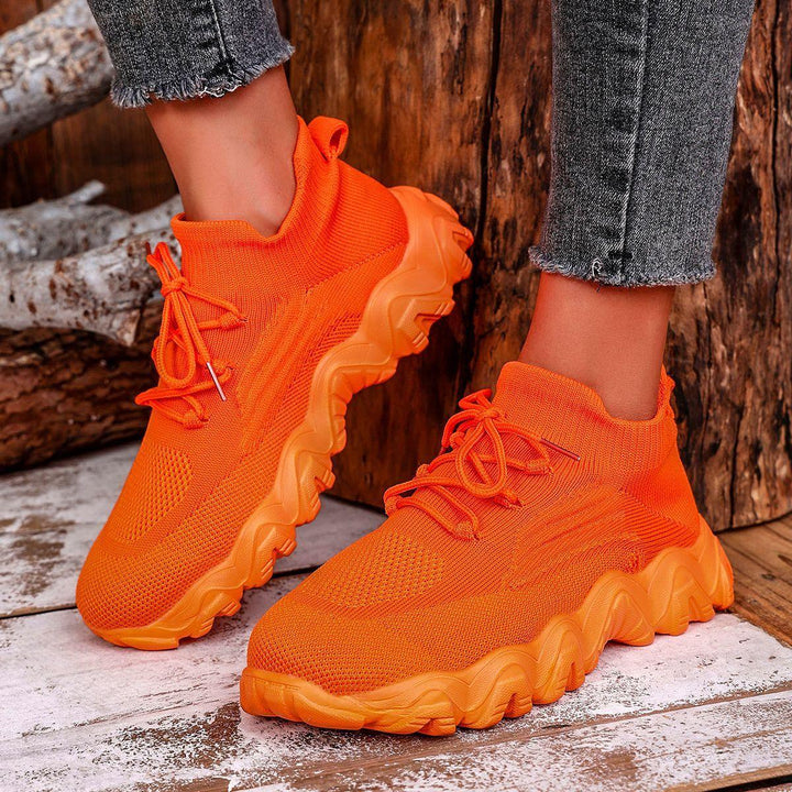 Bright orange knit sneakers with thick, wavy soles worn with frayed gray jeans on wooden floor