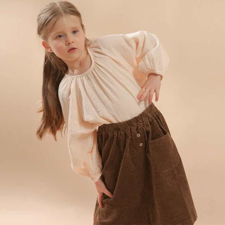 Young girl wearing a cream long-sleeve blouse and brown corduroy skirt against beige background