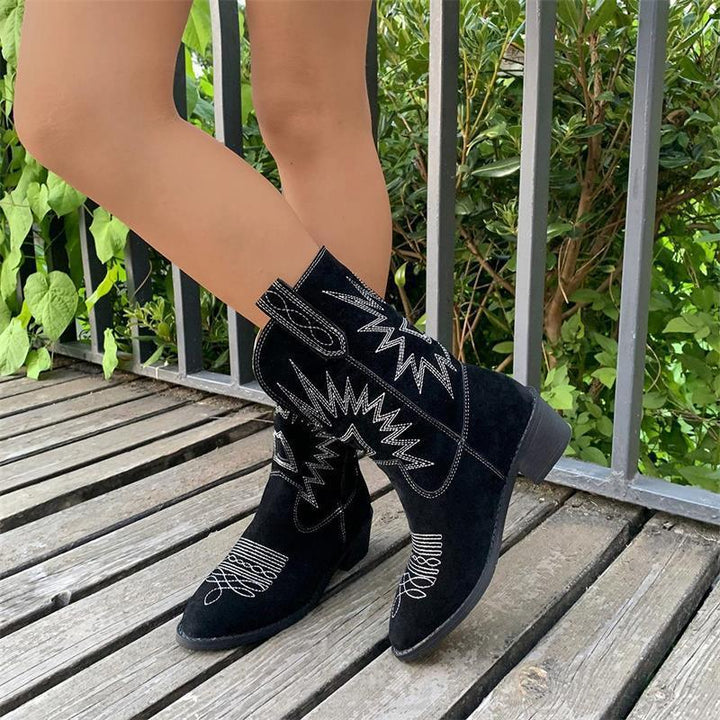 Close-up of black suede western ankle boots with white stitching worn on wooden deck near green foliage