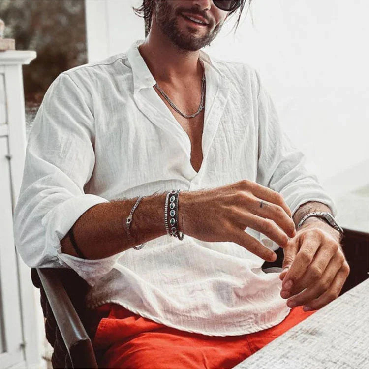 Man wearing white linen shirt, red pants, and multiple bracelets sitting at table with relaxed pose