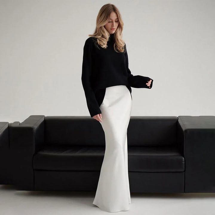 Woman wearing black sweater and long white skirt standing in front of black sofa in minimalist room