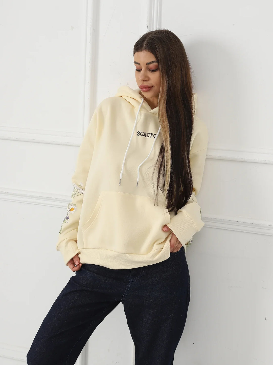 Woman wearing cream hoodie with floral sleeve embroidery and black jeans against white wall
