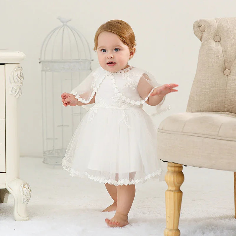 Toddler girl wearing a white lace dress standing barefoot in a bright room with white furniture