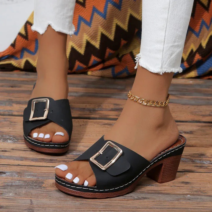 Black heeled sandals with large buckle detail worn with white jeans and gold ankle bracelet on wooden floor