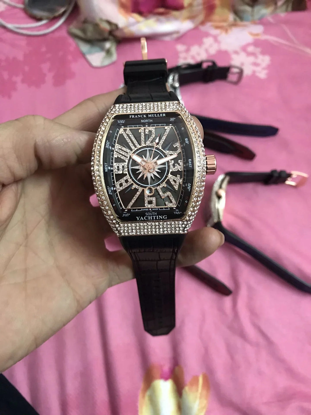 Close-up of a luxury Franck Muller Yachting watch with diamond bezel and black leather strap held over a pink floral background