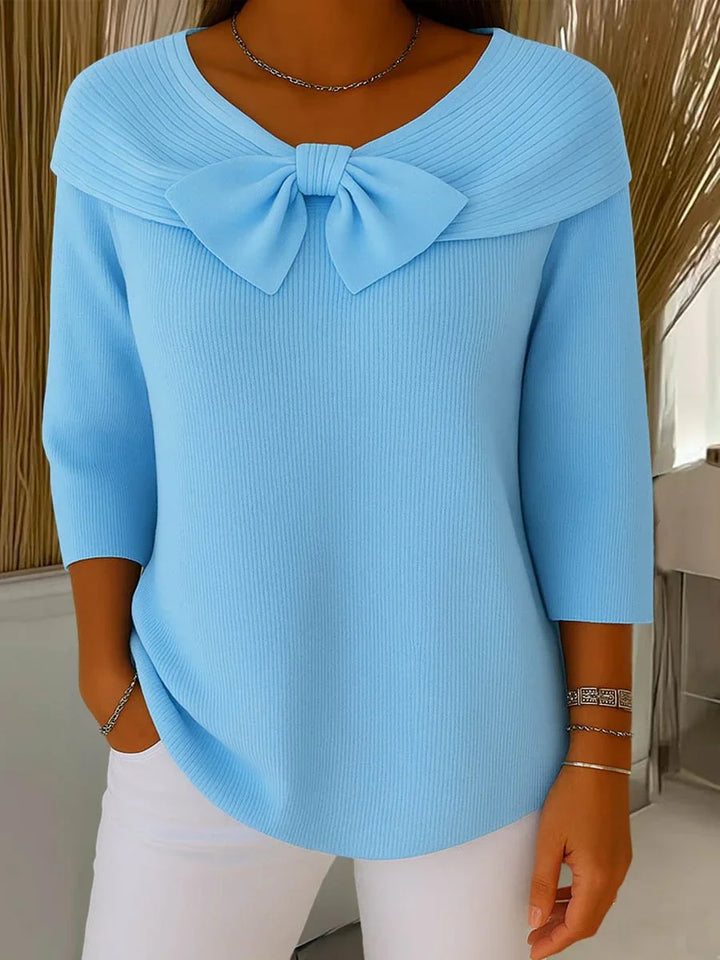 Woman wearing light blue ribbed sweater with large bow detail and 3/4 sleeves, paired with white pants