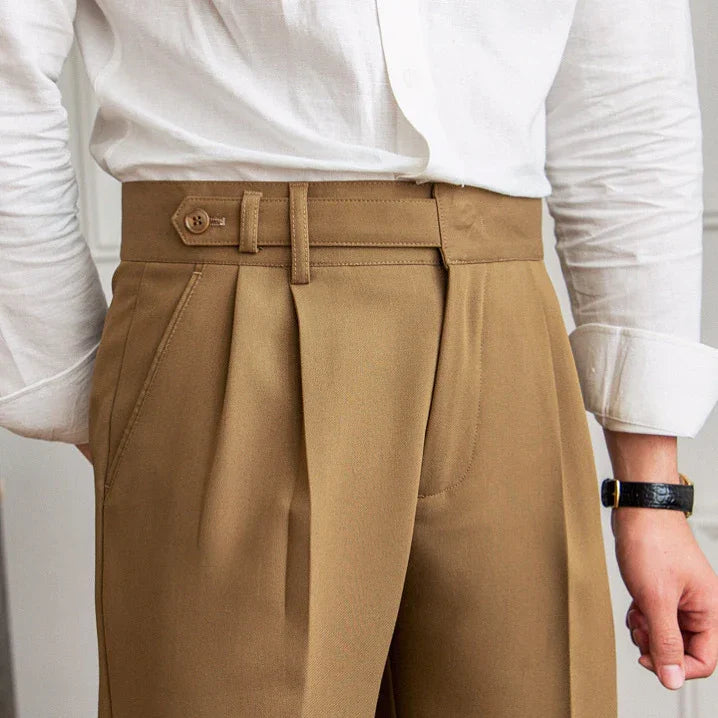 Close-up of man wearing high-waisted camel trousers with pleats and a buttoned waistband, paired with a white shirt.