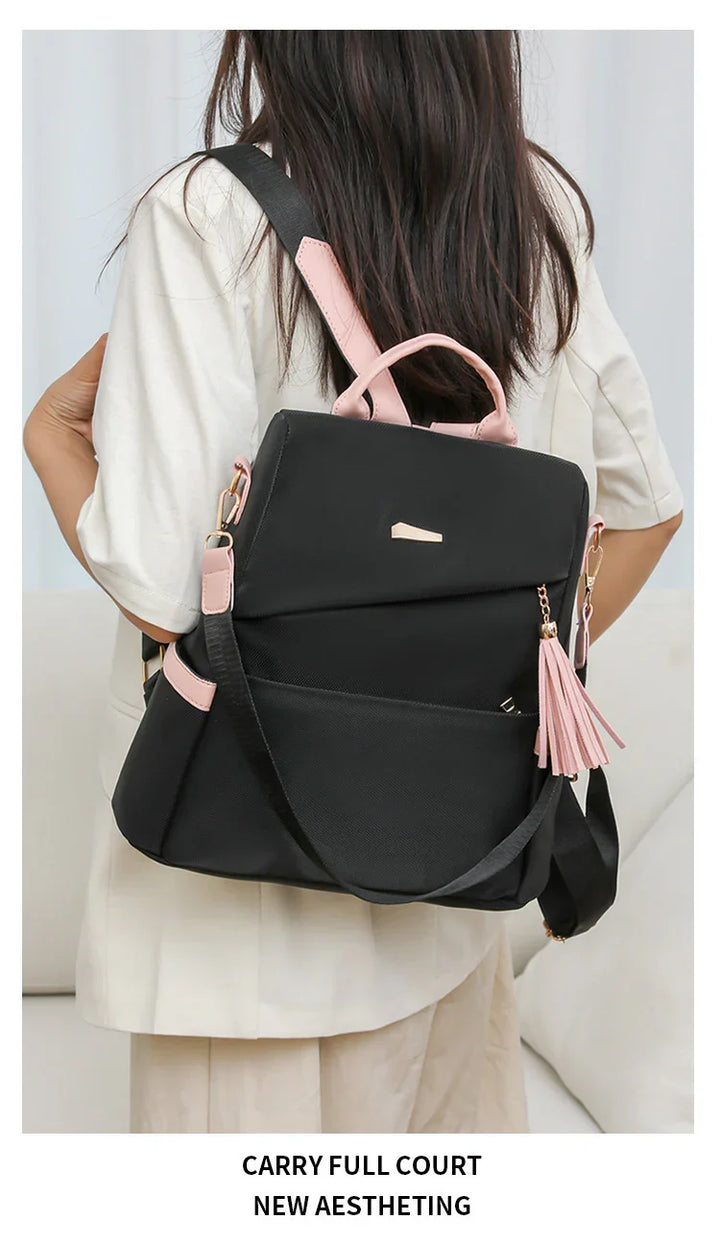 Black backpack with pink accents and tassel worn by person with long dark hair and beige outfit