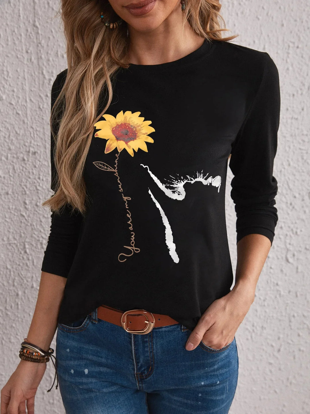 Woman wearing black long sleeve shirt with sunflower graphic and text You are my sunshine, paired with jeans and brown belt