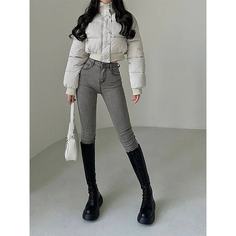 Woman wearing a white cropped puffer jacket, gray skinny jeans, black knee-high boots, holding white handbag