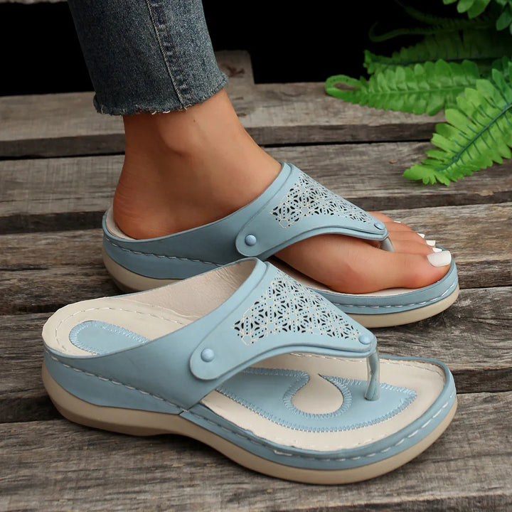 Pair of light blue leather thong sandals with floral cutout design worn by a person on wooden floor with green plant