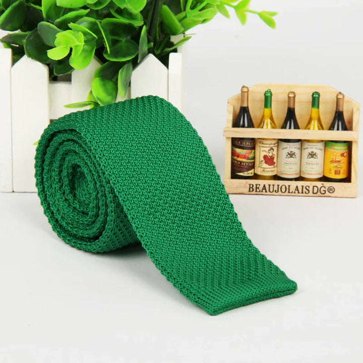 Green knitted slim tie displayed rolled near a small wooden crate with miniature wine bottles and green plant in white picket fence pot