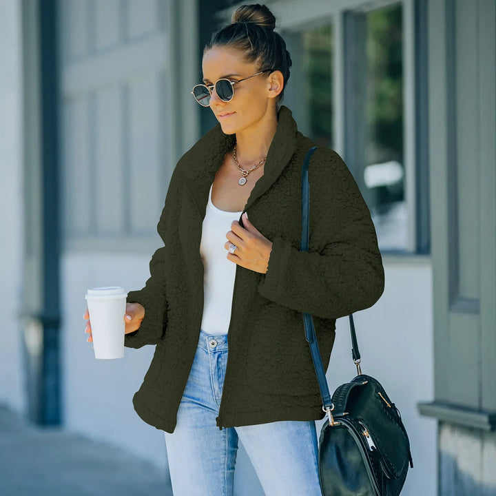 Woman wearing black sherpa fleece jacket with white tank top and jeans holding coffee cup outdoors
