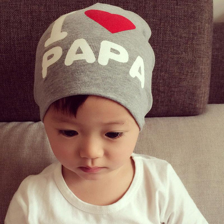 Toddler wearing grey beanie with 'I ❤️ PAPA' text and white shirt sitting on beige sofa