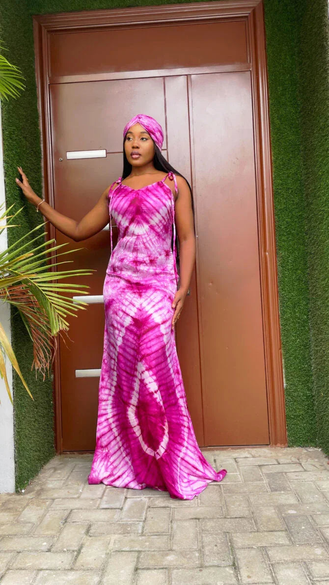 Woman in vibrant pink and white tie-dye maxi dress and matching headwrap standing by brown door with green foliage