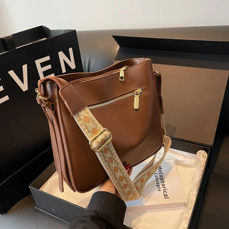 Brown leather crossbody bag with gold zippers and patterned adjustable strap inside black gift box