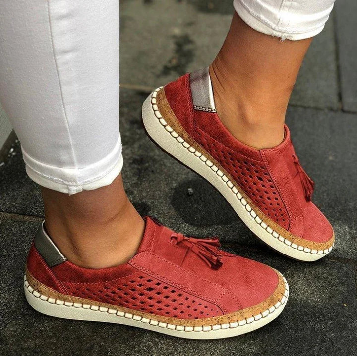 Close-up of red suede slip-on sneakers with tassels and perforated sides worn with white pants on black pavement