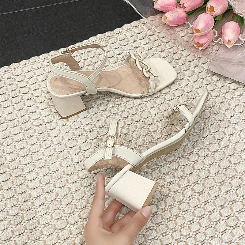 Pair of elegant off-white block heel sandals with ankle straps and chain detail on textured beige rug with pink tulips