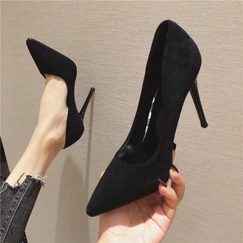 Black suede stiletto high heels worn and held by person against beige wall
