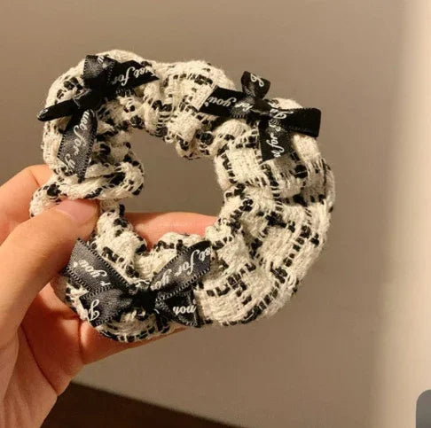 Hand holding a black and white tweed scrunchie with black ribbons adorned with white script text