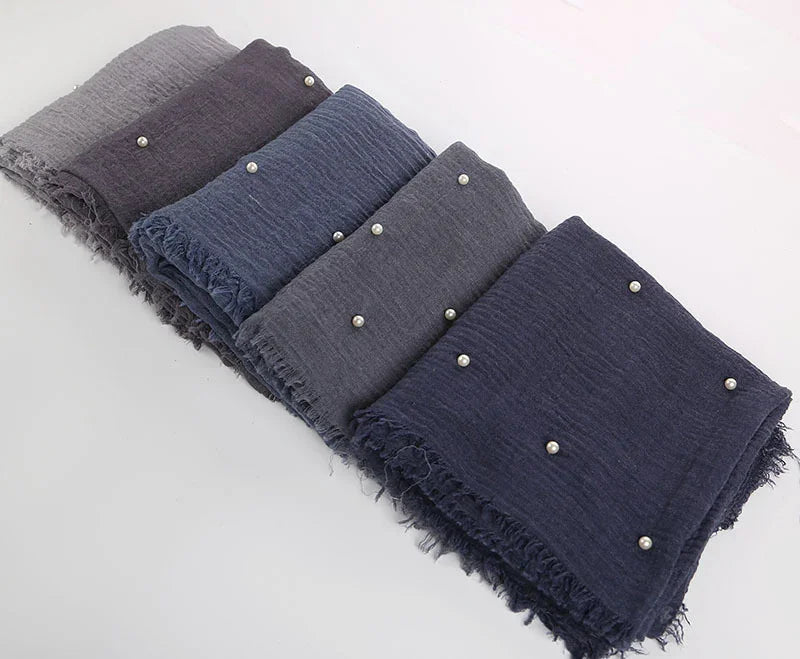 Folded textured scarves in shades of gray and blue with small pearl accents on a white surface
