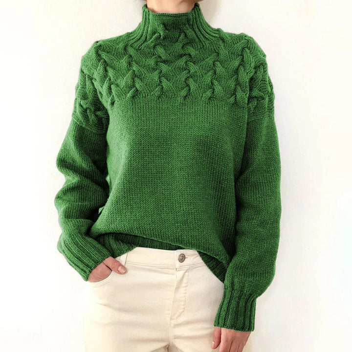 Person wearing a green cable knit turtleneck sweater with white pants against a plain background