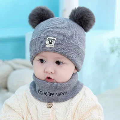Baby wearing gray knit hat with pom-poms and matching neck warmer with Love me more text