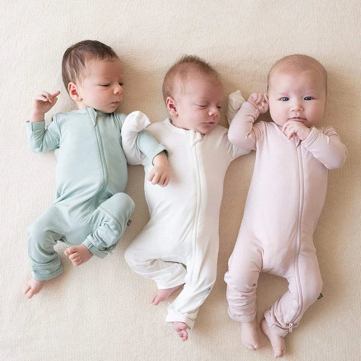 Three newborn babies lying side by side on a beige blanket wearing pastel-colored zip-up sleepwear