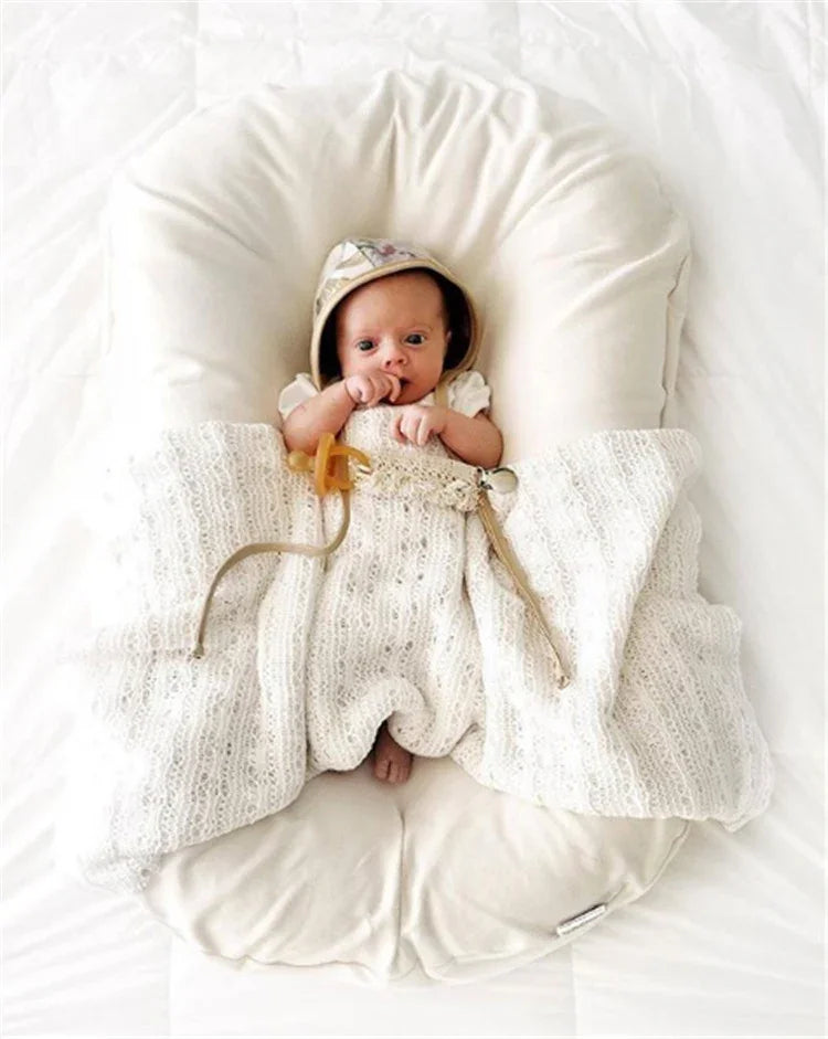 Newborn baby in a soft white knitted blanket and hood lying on a cushioned baby lounger