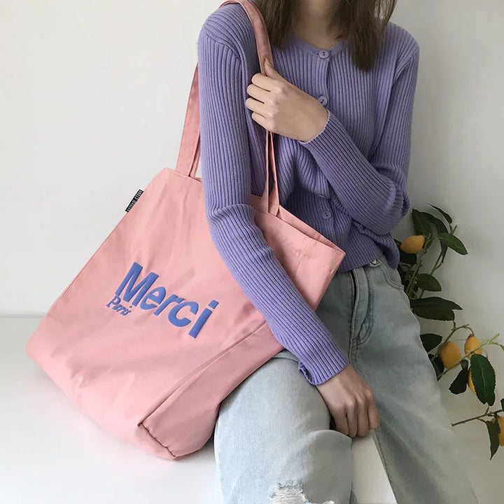 Woman wearing lavender cardigan and light jeans holding pink Merci Paris tote bag with lemon plant in background