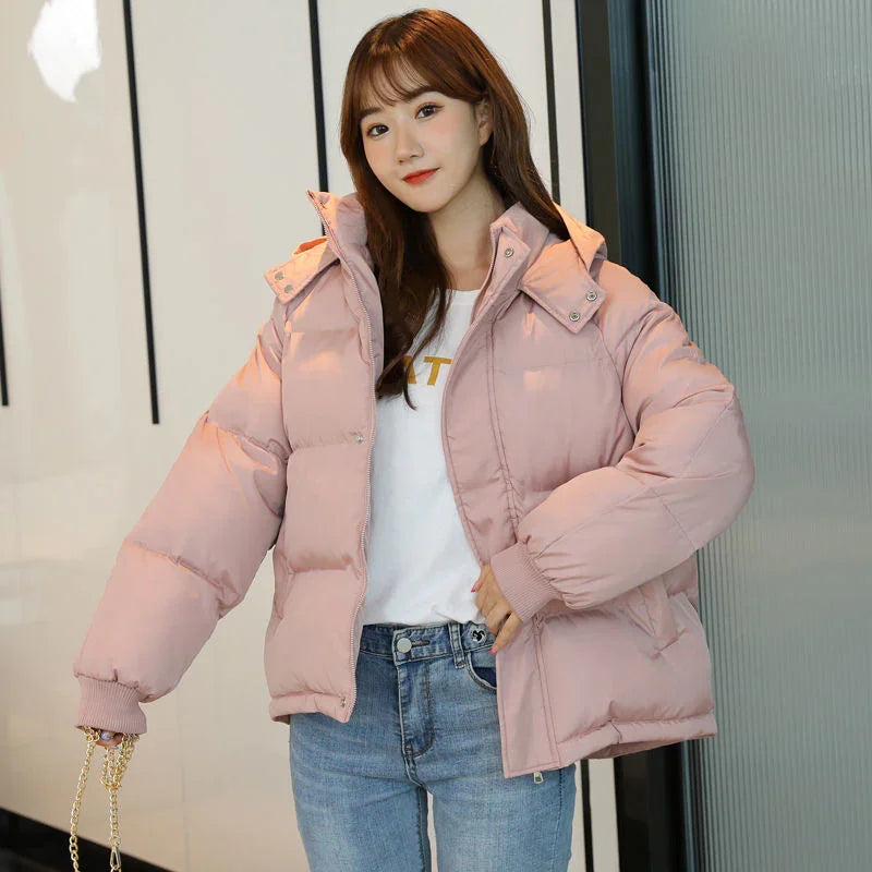 Young woman wearing a light pink puffer jacket with hood, white shirt, and blue jeans indoors