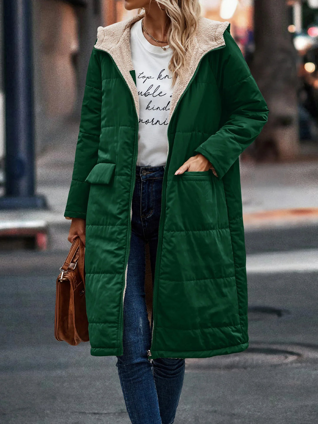 woman wearing long green quilted coat with sherpa lining, white printed t-shirt, blue jeans, and holding brown handbag on street