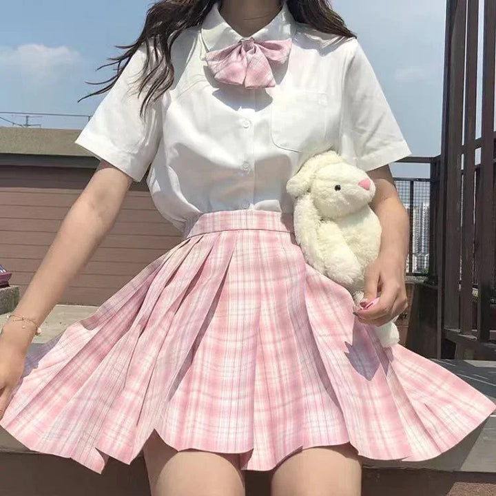 Woman in white blouse with pink bow and pink plaid pleated skirt holding plush bear outdoors
