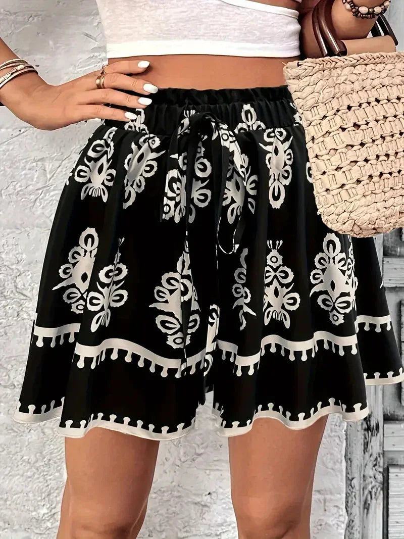 Woman wearing black and white patterned flowy skirt with elastic waist and tied bow, holding beige woven bag