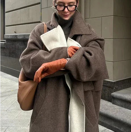 Stylish woman wearing oversized brown wool coat, tan leather gloves, glasses, and carrying tan shoulder bag on city sidewalk
