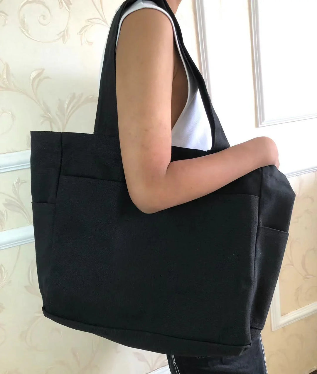Person holding a large black tote bag with wide straps over shoulder indoors