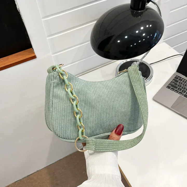 Green corduroy handbag with chain strap held over white desk with black lamp and laptop