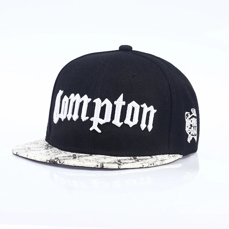 Black snapback hat with white embroidered Compton text and snakeskin pattern brim