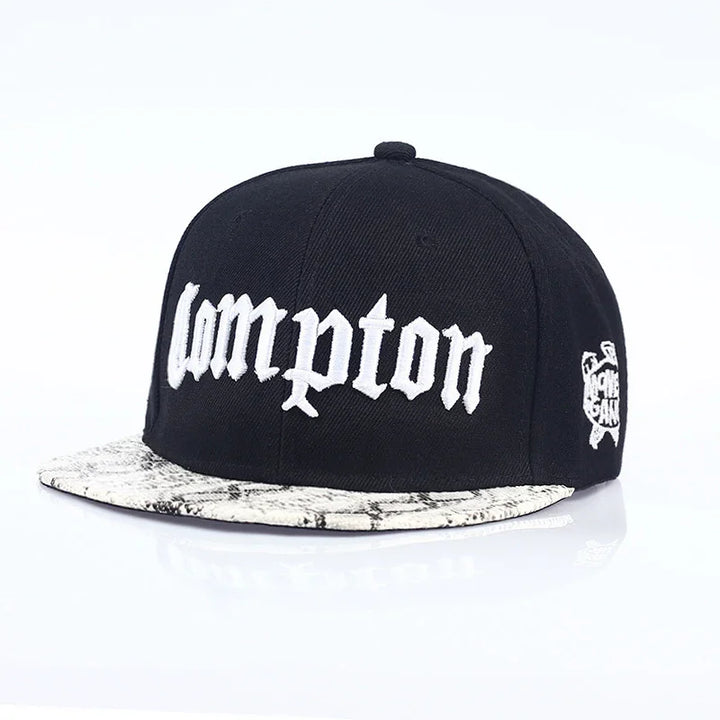 Black snapback hat with white embroidered Compton text and snakeskin pattern brim