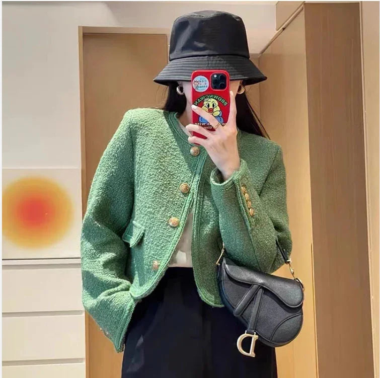 Woman wearing green textured jacket with gold buttons, black bucket hat, black saddlebag purse, taking mirror selfie