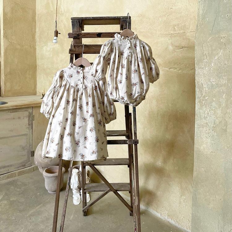 Two vintage-style cream floral baby dresses hanging on a rustic wooden ladder against textured beige wall