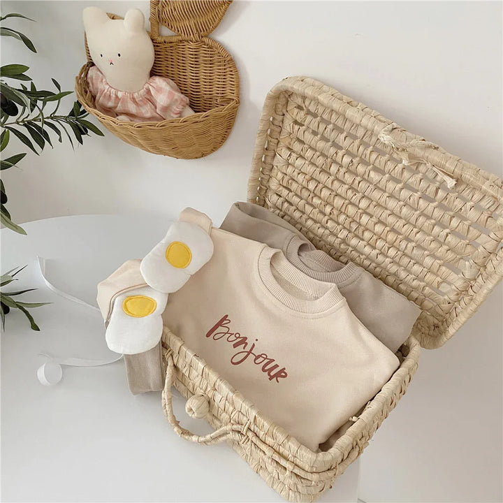 Wicker basket on white table holding beige and taupe sweaters, one with 'bonjour' text and fried egg mittens, plush bunny in wicker wall basket