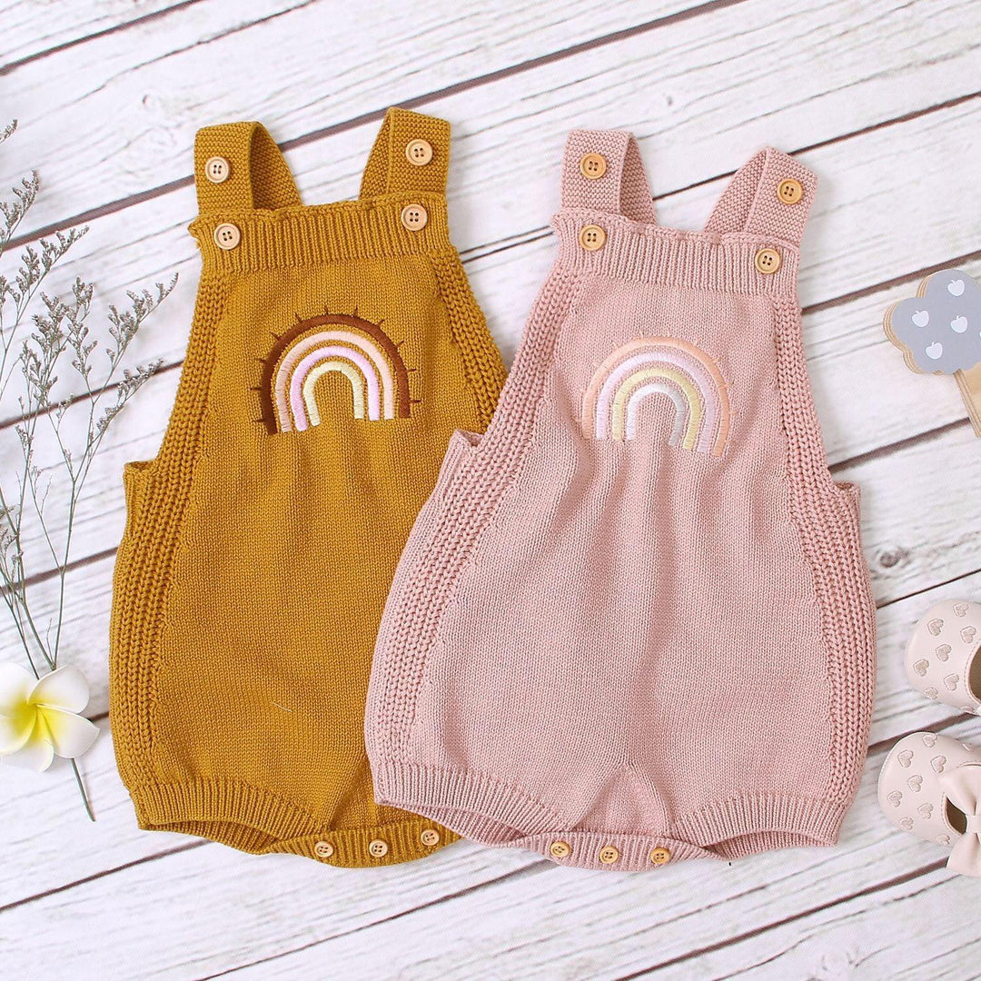 Two knitted baby rompers, mustard and blush pink, with rainbow embroidery on white wooden background