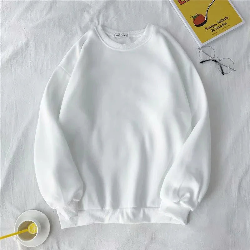 Plain white crewneck sweatshirt on white surface with glasses, book, and lemon drink