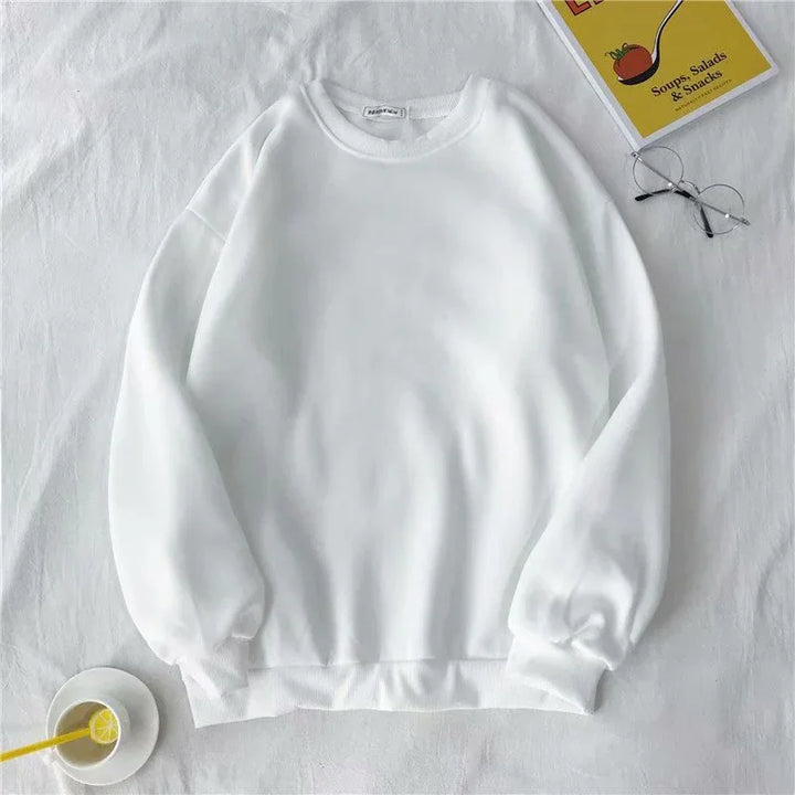 Plain white crewneck sweatshirt on white surface with glasses, book, and lemon drink