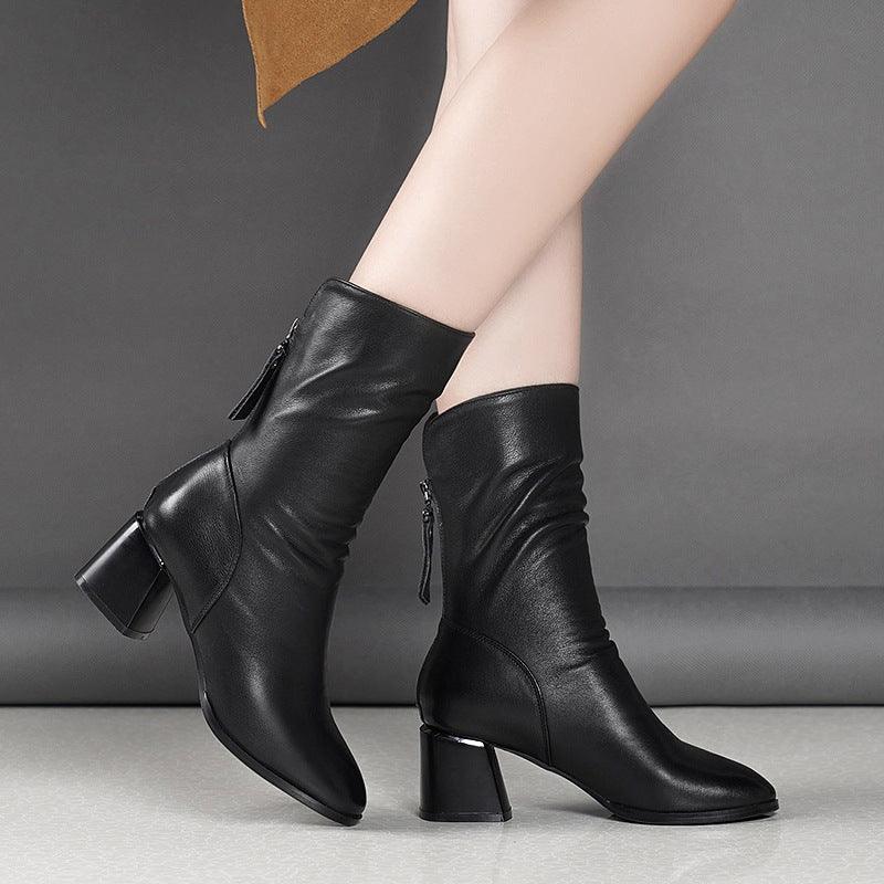 Pair of black leather mid-calf heeled boots worn on light-skinned legs against gray background